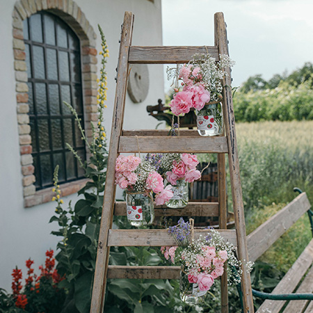 Eine Holzleiter vor einer Hauswand, auf der Blumenvasen mit Blumen aus Gläsern hängen.