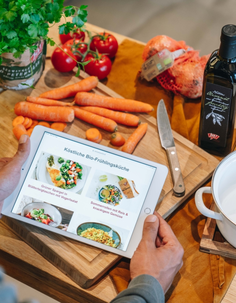 Tablet mit vier Bio-Frühstücksrezepten liegt auf einem Holzbrett, umgeben von frischem Karotten, Zwiebeln, Tomaten, Olivenöl und einem Messer. Eine Person hält das Tablet.
