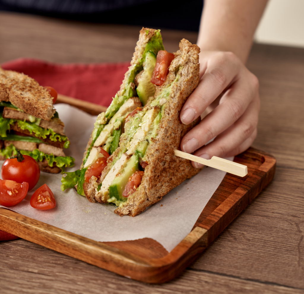 Jemand hält ein Bio-Clubsandwich in der Hand, welches auf einem Holzbrett liegt.