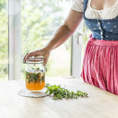 Jemand stellt ein Glas Oxymel auf den Tisch, daneben liegen Kräuter.