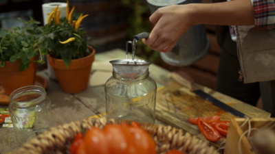 Jemand gießt Wasser durch ein Sieb mit Bio-Paradeisersamen, das auf einem großen Glas liegt.