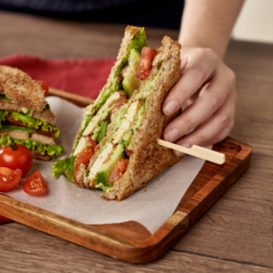 Jemand hält ein Bio-Clubsandwich in der Hand, welches auf einem Holzbrett liegt.