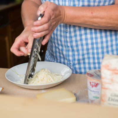 Jemand reibt Bio-Käse, daneben stehen Ja! Natürlich Bio-Schlagobers und Ja! Natürlich Bio-Dinkelmehl.