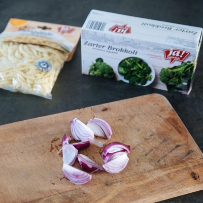Geschnittene Bio-Zwiebel auf einem Holzbrett, dahinter liegt eine Packung Ja! Natürlich Bio-Eierspätzle und eine Packung zarter Brokkoli.