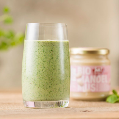 Ein Glas mit grünem Smoothie vor Ja! Natürlich Bio-Mandelmus.