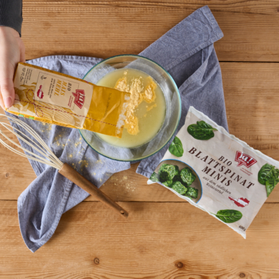 Jemand gibt Ja! Natürlich Bio-Maismehl in eine Schüssel. Daneben ist eine Packung Blattspinat.