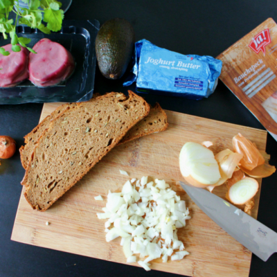Ein Holzbrett mit einer Scheibe Brot, Zwiebel und Ja! Natürlich Bio-Joghurtbutter und Bauchspeck.