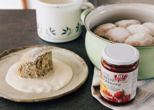 Buchteln auf Vanillesauce, daneben ein Glas Ja! Natürlich Bio-Ribisel-Erdbeermarmelade.