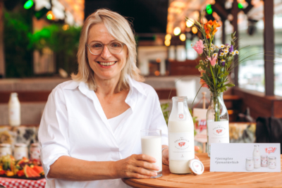 Martina Hörmer steht bei Holztisch und hält Glas Vollmilch von Ja! Natürlich in der Hand, daneben steht die Ja! Natürlich Vollmilch in der Glasflasche