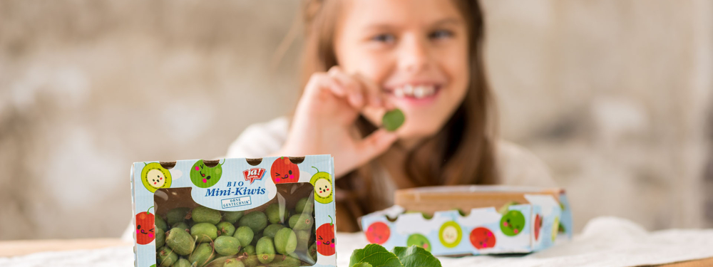 Packung Ja! Natürlich Bio-Mini-Kiwis im Vordergrund, Kind hält Kiwi im Hintergrund.