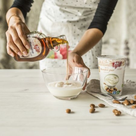 Jemand schüttet Ja! Natürlich Bio-Ahornsirup in eine Schale mit Mandeljoghurt.