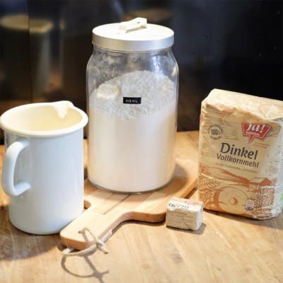 Ja! Natürlich Bio-Dinkel-Vollkornmehl und Ja! Natürlich Bio-Germ zusammen mit einem Vorratsglas voll Mehl und einem Krug Milch auf einem Holzbrett.