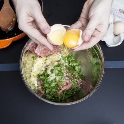 Jemand gibt ein Bio-Ei in eine Schüssel mit Zwiebel, gehackter Petersilie und Faschiertem.