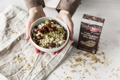 Jemand hält eine Schüssel mit Bio-Seewinkler Reis von Ja! Natürlich mit Bio-Cottage Cheese, Oliven und garniert mit Kräutern.