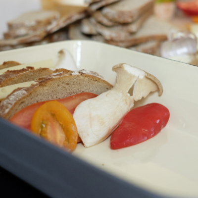 Eine Backform mit einem Bio-Kräuterseitling, halbierten Paradeiser und drei Brotscheiben mit Bergkäse dazwischen.