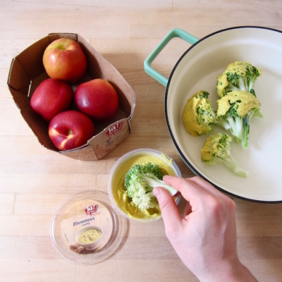 Jemand taucht Bio-Brokkoli in Ja! Natürlich Bio-Curry Hummus. Daneben steht ein Karton mit Ja! Natürlich Bio-Äpfeln.