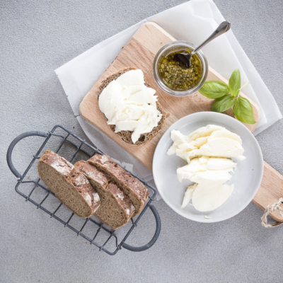 Auf einem Holzbrett liegt ein Brot mit Bio-Mozzarella, ein Glas mit grünem Pesto und ein Teller mit geschnittenem Mozzarella.