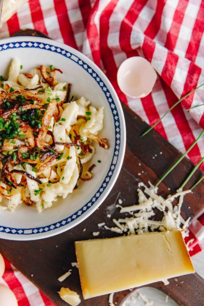 Teller mit Käsespätzle, garniert mit Röstzwiebeln und Schnittlauch, auf Holzbrett mit Käse und Eierschale daneben.