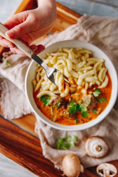 Gabel mit Spätzle über einem Teller Geschnetzeltem mit Spätzle und Kräutern, auf einem Holztisch mit Pilzen im Vordergrund.