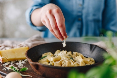 Jemand streut Bio-Bergkäse über Tortelloni in einer Gusseisenpfanne.