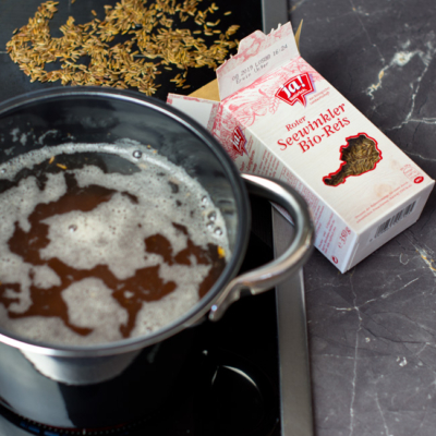 Ein Topf mit kochendem Seewinkler Bio-Reis, daneben eine ausgeleerte Packung Ja! Natürlich Seewinkler Bio-Reis.