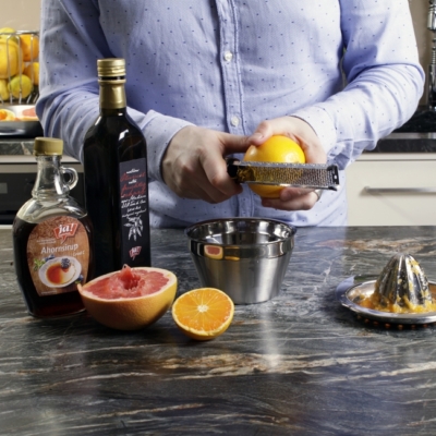 Jemand reibt Blutorangen Zeste ab. Daneben steht eine Flasche Ja! Natürlich Bio-Ahornsirup und Aceto Balsamico.