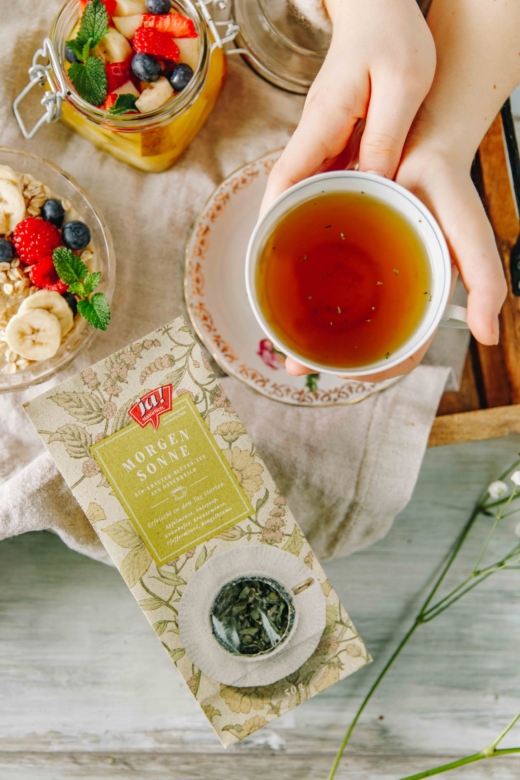 Jemand hält eine Tasse Tee. Daneben liegt eine Packung Ja! Natürlich Bio-Morgensonne Tee.