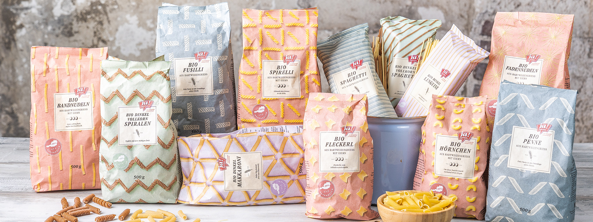 Bio-Bandnudeln, Bio-Dinkel Vollkorn Spiralen, Bio-Fusilli, Bio-Spirelli, Bio-Dinkel Makkaroni, , Bio-Dinkel-Vollkorn Spaghetti, Bio-Spaghetti, Bio-Dinkel Linguine, Bio-Fleckerl, Bio-Hörnchen, Bio-Fadennudeln und Bio-Penne von Ja! Natürlich sind in der Papierverpackung auf einem Tisch aufgestellt.