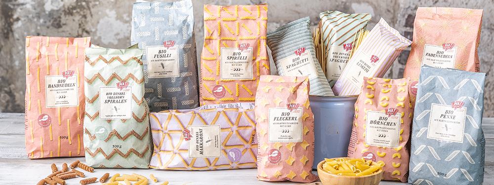 Bio-Bandnudeln, Bio-Dinkel Vollkorn Spiralen, Bio-Fusilli, Bio-Spirelli, Bio-Dinkel Makkaroni, , Bio-Dinkel-Vollkorn Spaghetti, Bio-Spaghetti, Bio-Dinkel Linguine, Bio-Fleckerl, Bio-Hörnchen, Bio-Fadennudeln und Bio-Penne von Ja! Natürlich sind in der Papierverpackung auf einem Tisch aufgestellt.