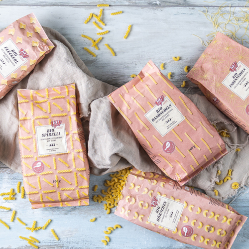 Bio-Fleckerl, Bio-Spirelli, Bio-Bandnudeln, Bio-Hörnchen und Bio-Fadennudeln von Ja! Natürlich in der Papierverpackung liegen auf einem Tisch