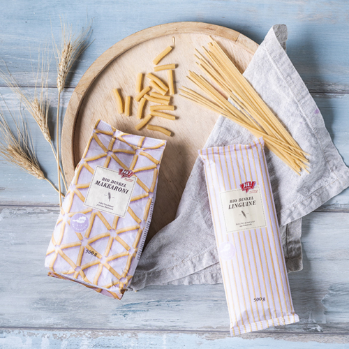 Bio-Dinkel Makkaroni und Bio-Dinkel Linguine von Ja! Natürlich liegen in der Papierverpackung auf einem Holzbrett