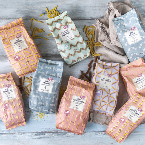 Bio-Fleckerl, Bio-Spirelli, Bio-Fusilli, Bio-Dinkel-Vollkorn Spiralen, Bio-Bandnudeln, Bio-Dinkel-Makkaroni, Bio-Penne, Bio-Hörnchen, Bio-Fadennudeln in Papierverpackung von Ja! Natürlich liegen auf einem Tisch