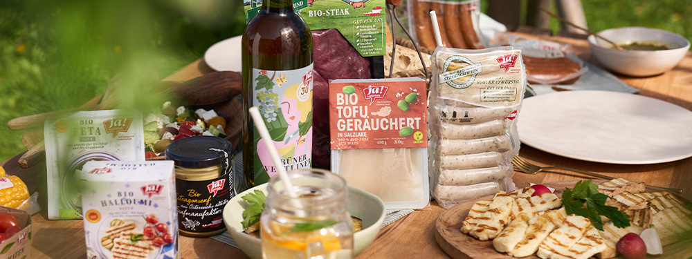 Auf einem Tisch stehen Grillprodukte, wie Fleisch, Würstel, Gemüse, Salat, Grillkäse und Getränke wie Wein und Limonade