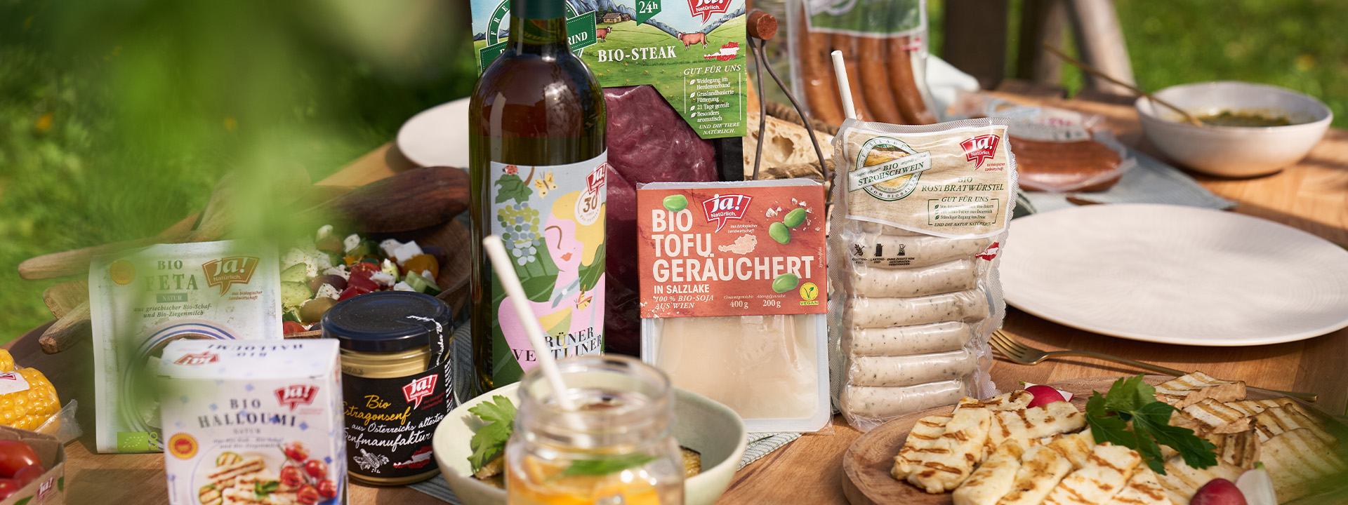 Auf einem Tisch stehen Grillprodukte, wie Fleisch, Würstel, Gemüse, Salat, Grillkäse und Getränke wie Wein und Limonade