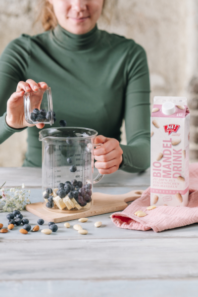 Jemand gibt die Zutaten für einen Smoothie in einen Messbecher, daneben ein Karton Ja! Natürlich Bio-Mandel-Drink.