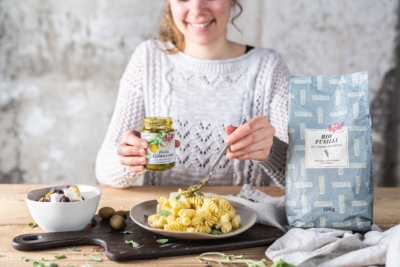 Jemand gibt einen Löffel Ja! Natürlich Bio-Pesto über einen Teller mit Fusilli.