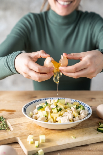 Jemand schlägt ein Bio-Ei über Semmelstücke, Erdäpfel und Zucchini.