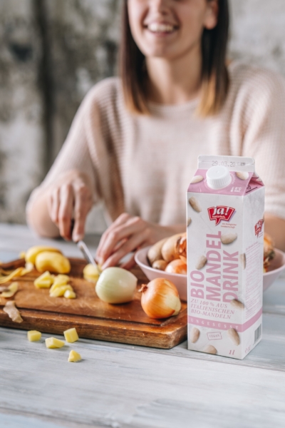 Jemand schneidet Bio-Erdäpfel auf einem Brett, daneben steht ein Tetrapak Ja! Natürlich Bio-Mandel Drink.