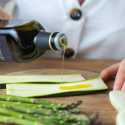 Bio-Olivenöl wird auf eine aufgeschnittene Zucchini gegossen, daneben liegen Spargel und Zwiebel.