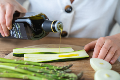 Bio-Olivenöl wird auf eine aufgeschnittene Zucchini gegossen, daneben liegen Spargel und Zwiebel.