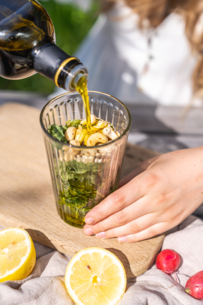 Jemand schüttet Bio-Olivenöl über ein Glas mit Cashewkernen, Grün von Radieschen und Knoblauch.