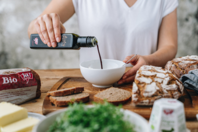 Jemand gießt Ja! Natürlich Bio-Kürbiskernöl in eine Schüssel, Brot und Schafkäse liegen daneben.