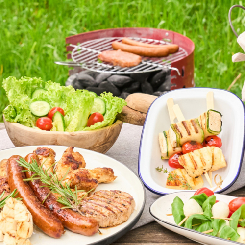 Vielfältige Grillspezialitäten von Ja! Natürlich, angerichtet auf Tellern und im Hintergrund ein Griller mit Bio-Würsteln.