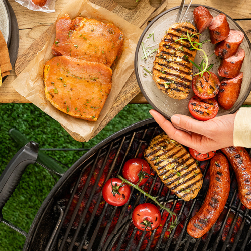 Bio-Fleisch und Bio-Gemüse auf dem Rost eines Grills, daneben auf einem Tisch rohes und frisch mariniertes Bio-Fleisch.