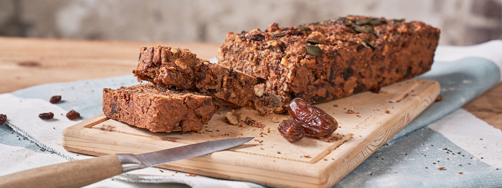 Veganer Bio-Früchtestollen frisch zubereitet auf einem Holzbrett, daneben Bio-Datteln und ein Küchenmesser.