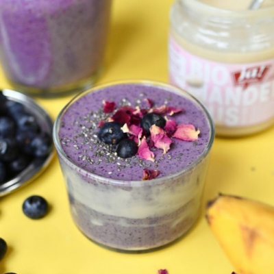 Der fertige Blueberry Almond Protein Lassi in einem Gals, garniert mit frischen Bio-Heidelbeeren, Rosenblüten, Chiasamen und gehakten Bio-Pistazien.