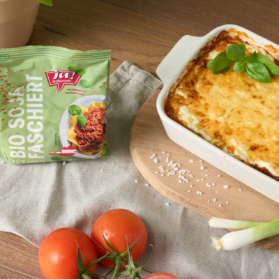 Die fertige High Protein Lasagne steht auf einem Tisch und daneben eine Packung des Bio-Soja-Faschiert und Bio-Paradeiser.