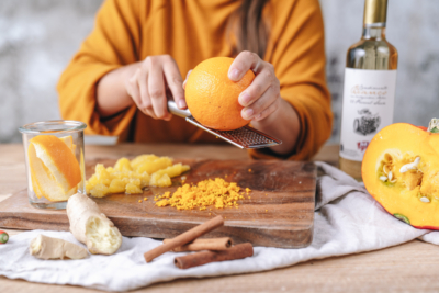 Jemand reibt die Schale einer Orange auf ein Brett. Daneben steht eine Flasche Ja! Natürlich Bio-Condimento Bianco.