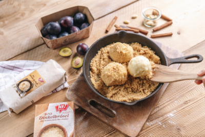 Jemand wälzt Knödel in Brösel, daneben liegt eine Packung Ja! Natürlich Bio-Dinkelgrieß.