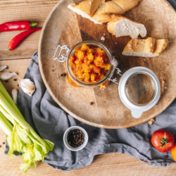 Bio-Paradeiser-Stangensellerie-Chutney auf einem Brett mit Baguette und Gemüse.